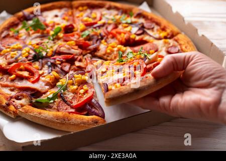 La main caucasienne humaine prend une tranche de grande pizza de la boîte de carton sur la table en bois blanc Banque D'Images