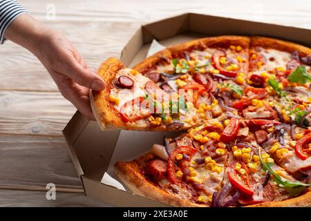 La main caucasienne humaine prend une tranche de grande pizza de la boîte de carton sur la table en bois blanc Banque D'Images