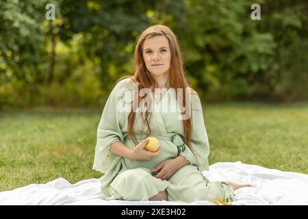 Blanche jolie femme enceinte est assise sur une couverture, tenant, mangeant pomme dans le parc. Profiter de la prairie, détente. Mode de vie sain. Accouchement Banque D'Images