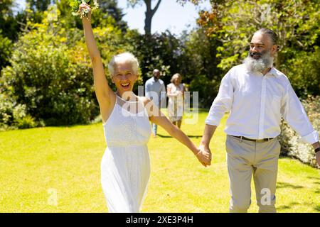 Célébrant à l'extérieur, couple senior souriant, femme tenant le bouquet, homme tenant sa main Banque D'Images