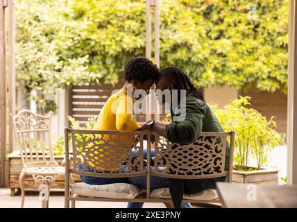 Couple afro-américain assis sur le banc, partageant un moment intime, souriant en plein air Banque D'Images