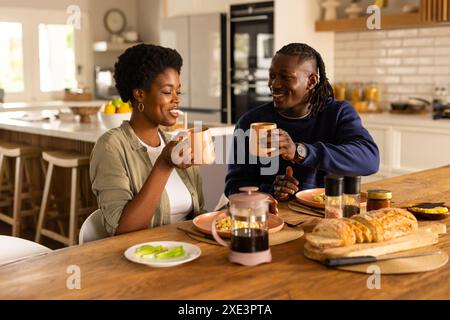 À la maison, couple profitant du petit déjeuner ensemble, souriant et tenant des tasses à café dans la cuisine Banque D'Images