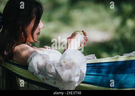 Belle fille mangeant une pomme s'amusant dans un hamac de parc, une journée ensoleillée capturant l'amour et le bonheur. Banque D'Images