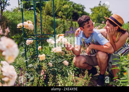 Heureux jeune couple appréciant les fleurs ensemble dans le jardin d'été. Jardiniers vérifiant les plantes portant des tabliers dans le jardin de chalet rose Banque D'Images