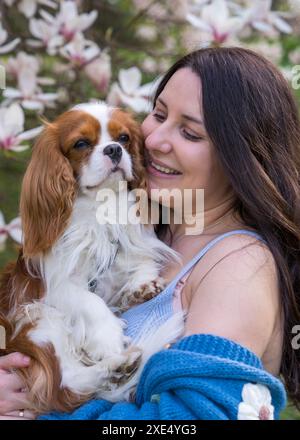 Une femme mignonne embrasse son beau chien, un épaniel cocker, dans la rue, dans le parc, près d'un magnolia. Cavalier King Charles Cocker Spaniel. Close-u Banque D'Images