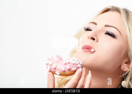 Femme blonde apprécie un beignet givré rose avec des guimauves, se livrant à ses saveurs sucrées et tentantes. Son expression véhicule la satisfaction pure et Banque D'Images