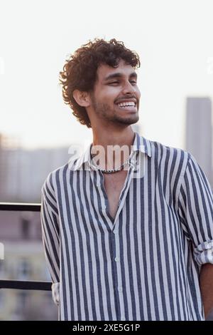 Gros plan d'un portrait masculin d'un jeune homme de race mixte. Un gars attrayant aux cheveux bouclés dans une chemise rayée sourit contre le b. Banque D'Images