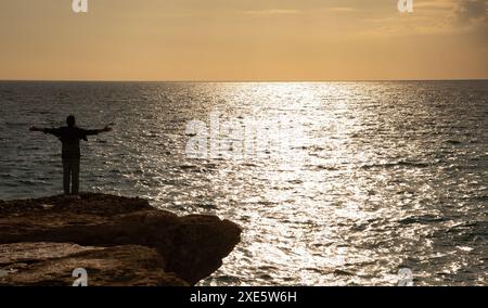 Silhouette d'homme levant les mains ou les bras ouverts au coucher du soleil au bord de la falaise dans l'océan Banque D'Images