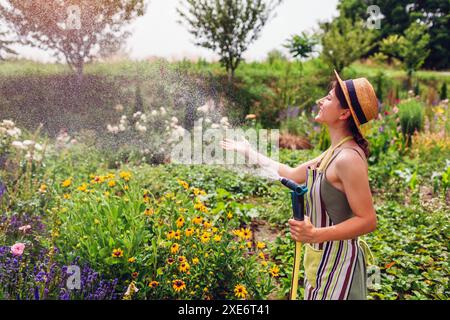 Jardinier de femme joyeuse dans le chapeau de paille tenant le tuyau de jardin ayant le plaisir d'arroser les plantes dans le jardin d'été. Prendre soin des fleurs à la ferme Banque D'Images