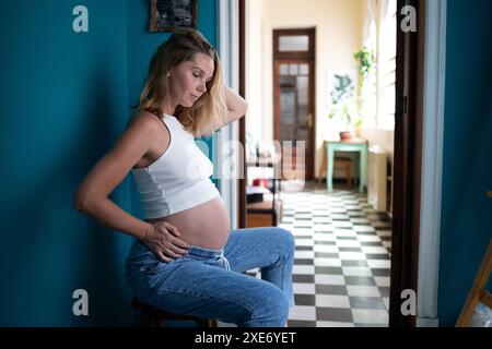 Une femme enceinte en vêtements décontractés s'assoit pensivement, main sur le ventre, dans une maison confortable Banque D'Images