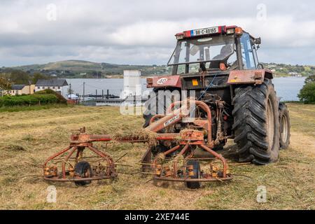 Un agriculteur basé à Schull tourne l'herbe coupée pour faire du foin à l'aide d'un tracteur case 4230 avec Schull Harbour, West Cork, Irlande comme toile de fond. Banque D'Images