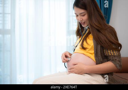 Femme enceinte asiatique écoutant le rythme cardiaque du bébé avec stéthoscope à la maison Banque D'Images