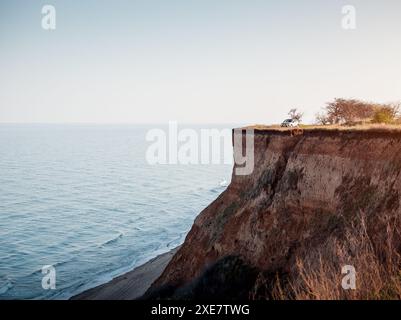 Voiture argentée garée au sommet d'un cllif au bord de la mer au coucher du soleil. Côte falsifiée de la mer Noire, sol sablonneux glissant lentement dans l'eau en raison de l'abrasion. Banque D'Images