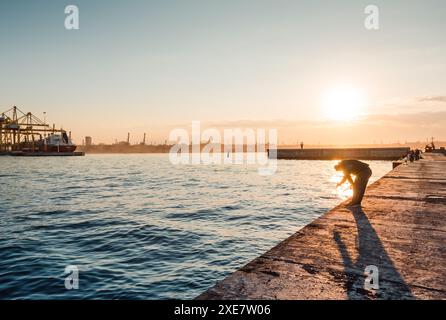 Jetée près du port de mer au coucher du soleil. Soleil couchant bas sur la baie et silhouettes de pêcheurs. Cargo amarré à gauche en cours de chargement. Banque D'Images