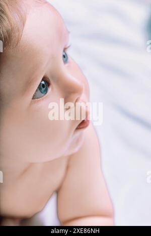 Bébé garçon avec de grands yeux bleus se trouve paisiblement sur un lit, regardant avec émerveillement et innocence. L'image capture la beauté de l'enfance et la joie parentale dans ainsi Banque D'Images