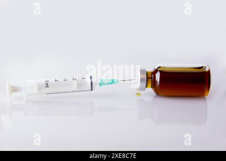 Seringue médicale avec aiguille coincée dans une bouteille de médicament injectable. Il y a une goutte sur la table. Fond blanc. Banque D'Images