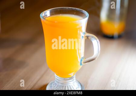 Un mug en verre transparent rempli de jus d'orange vif placé sur une table en bois. Le fond est flou avec un autre verre partiellement visible. Banque D'Images