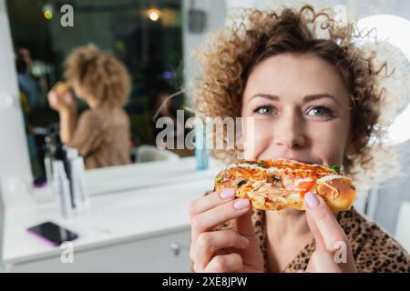 Jeune femme bouclée mangeant de la pizza dans un salon de beauté et souriante Banque D'Images