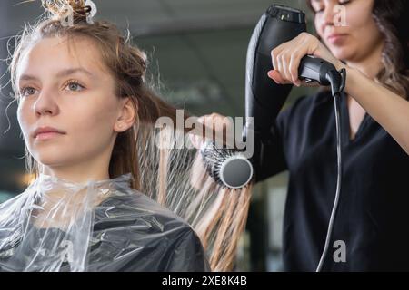 Gros plan des mains de coiffeurs séchant de longs cheveux blonds avec sèche-cheveux et brosse ronde Banque D'Images