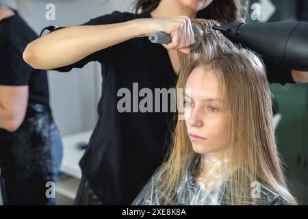 Gros plan des mains de coiffeurs séchant de longs cheveux blonds avec sèche-cheveux et brosse ronde Banque D'Images