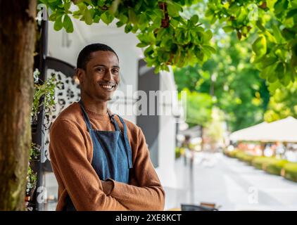 Jeune homme souriant d'origine marocaine dans un tablier de travail décontracté dans un restaurant en plein air. Il se tient confiant, les bras croisés, entouré de verdure. Banque D'Images