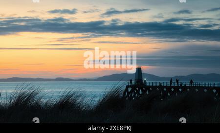 Alors que le soleil plonge sous l’horizon à Maryport, Cumbria, le ciel du coucher de soleil accentue parfaitement les silhouettes des pêcheurs et de l’emblématique jetée Banque D'Images