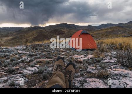 pieds d'homme avec des bottes à l'intérieur d'une tente orange dans les montagnes dans un coucher de soleil entouré de rochers et de végétation jaune en hiver dans la montagne des andes Banque D'Images