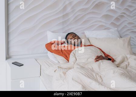 Homme afro-américain en chemise orange dort paisiblement dans un lit bien fait avec une literie blanche, mettant en valeur la détente et le confort. Téléphone avec réveil est sur la table de chevet à côté. Banque D'Images