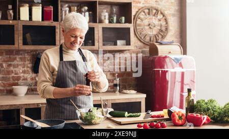 Femme senior ajoutant du poivre à la salade fraîche dans la cuisine à la maison Banque D'Images