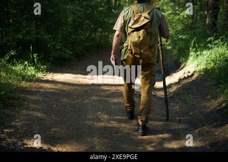 Vue arrière photo de coupe d'un forestier masculin méconnaissable portant un sac à dos tenant une tablette numérique et un bâton en bois marchant le long de la route Banque D'Images