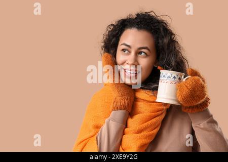 Belle jeune femme afro-américaine dans des mitaines chaudes avec tasse de vin chaud chaud sur fond brun Banque D'Images