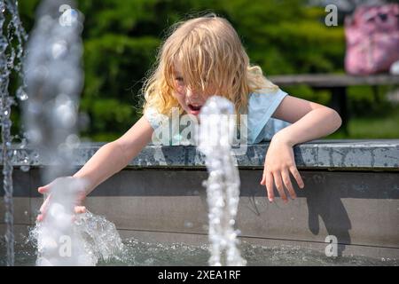 fille en jeans et une chemise bleu clair se tient près de la fontaine, laissant la brume d'eau refroidir son visage. Arbres verts et tables vides d'un cadre de foire de ville t Banque D'Images