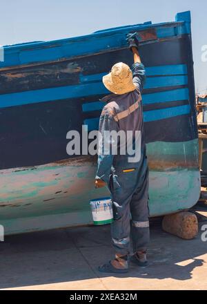 Homme marocain local face à l'extérieur peignant un bateau bleu au soleil, au port en été, authentique scène portuaire franche à tous les jours, Essaouira, Maroc Banque D'Images