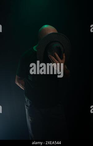 Homme mystérieux tenant un chapeau devant son visage posant pour la caméra. Portrait de studio. Fond sombre avec fumée artificielle. Banque D'Images