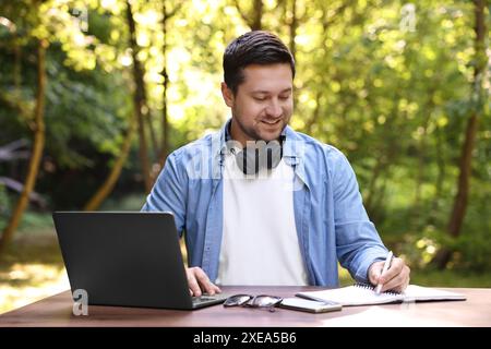 Souriant freelance travaillant avec un ordinateur portable et écrivant quelque chose à table à l'extérieur. Tâche distante Banque D'Images