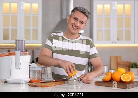 Homme souriant pressant l'orange fraîche avec presse-agrumes à la table en marbre blanc dans la cuisine Banque D'Images