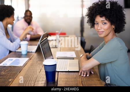 Souriant à la caméra, femme d'affaires avec ordinateur portable au bureau, collègues discutant en arrière-plan Banque D'Images