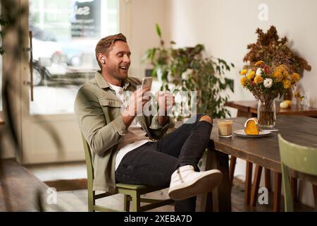 Portrait d'heureux beau jeune homme, assis dans un café avec téléphone portable, portant des écouteurs sans fil, écoutant de la musique ou podc Banque D'Images