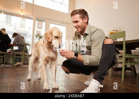 Portrait d'homme beau souriant avec son chien, assis sur le sol dans un café avec Golden retriever, donnant un régal Banque D'Images