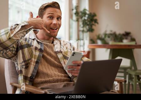 Portrait de drôle beau visiteur de café, jeune homme avec ordinateur portable et smartphone, montrant le signe de main de téléphone portable et souriant à la caméra Banque D'Images