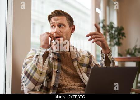 Portrait d'homme d'affaires sérieux parlant au téléphone, assis dans une chaise dans un café avec ordinateur portable, travaillant sur le projet, discutant w Banque D'Images