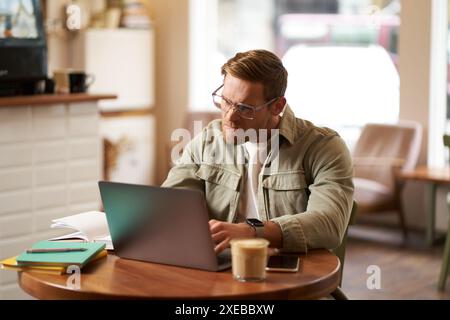 Portrait d'homme d'affaires dans des lunettes, est assis dans un café avec ordinateur portable, regarde concentré à l'écran avec sa tâche de projet, travaille à distance Banque D'Images