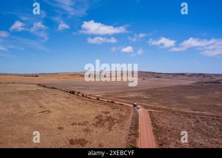 Vue aérienne d'une voiture et d'une caravane conduisant sur un chemin de terre à travers de vastes champs ouverts sous un ciel bleu clair Banque D'Images