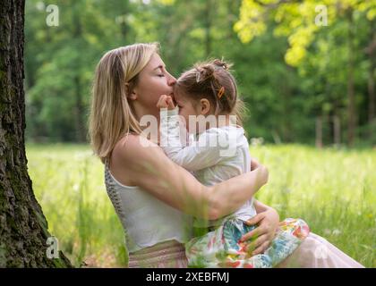 Maman et fille dans le parc. Maman est assise près d'un arbre et embrasse doucement sa petite fille. Journée ensoleillée, hautes herbes. Amour, soins. Maman et enfant. Banque D'Images