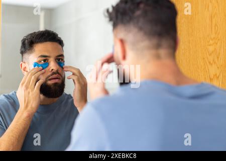 Homme appliquant des patchs sous les yeux tout en regardant dans le miroir de la salle de bain à la maison Banque D'Images