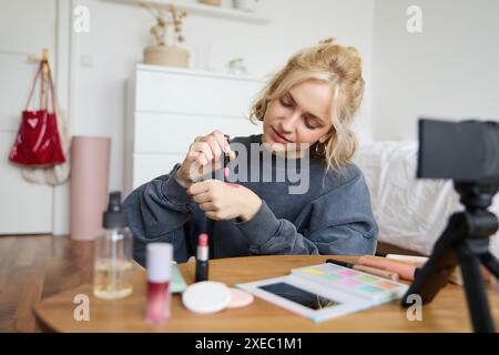 Femme blogueuse de beauté, enregistre la vidéo d'elle-même assise dans une pièce et évaluant les produits de maquillage, met sur le maquillage, tient le rouge à lèvres et Banque D'Images
