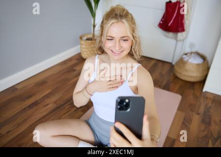 Portrait de jeune belle femme blonde, instructeur de fitness faisant des exercices à la maison sur tapis de yoga, prenant des selfies sur smartphone, R Banque D'Images