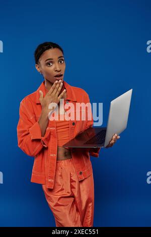 Jeune femme afro-américaine en tenue orange tient en toute confiance un ordinateur portable sur un fond bleu vif. Banque D'Images