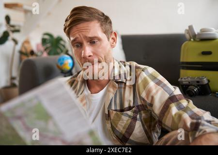 Portrait de l'homme regardant confus à la carte de voyage, choisir l'itinéraire, partir en vacances, préparer les vacances, assis dans le salon Banque D'Images