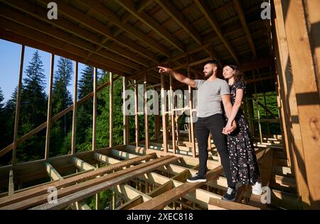 Homme et femme inspectant leur future habitation à ossature en bois nichée dans les montagnes près de la forêt. Jeune couple au chantier de construction tôt le matin. Concept de construction écologique contemporaine Banque D'Images